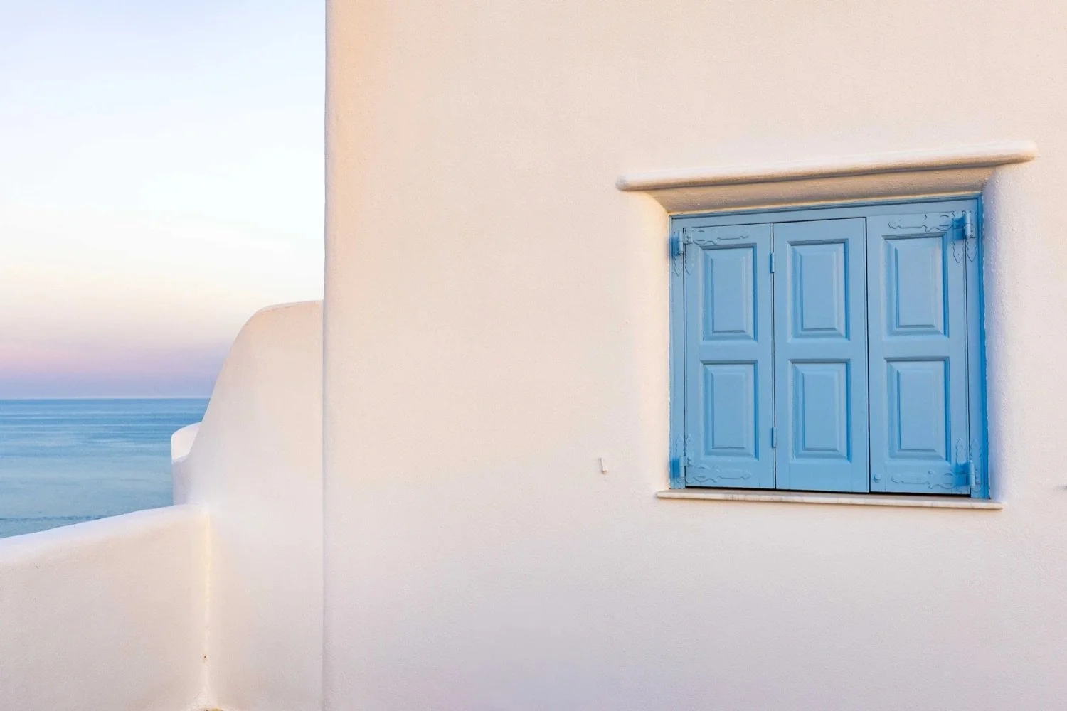 White building with a closed blue window on the right, overlooking the ocean at sunset or sunrise in a scenic coastal setting.