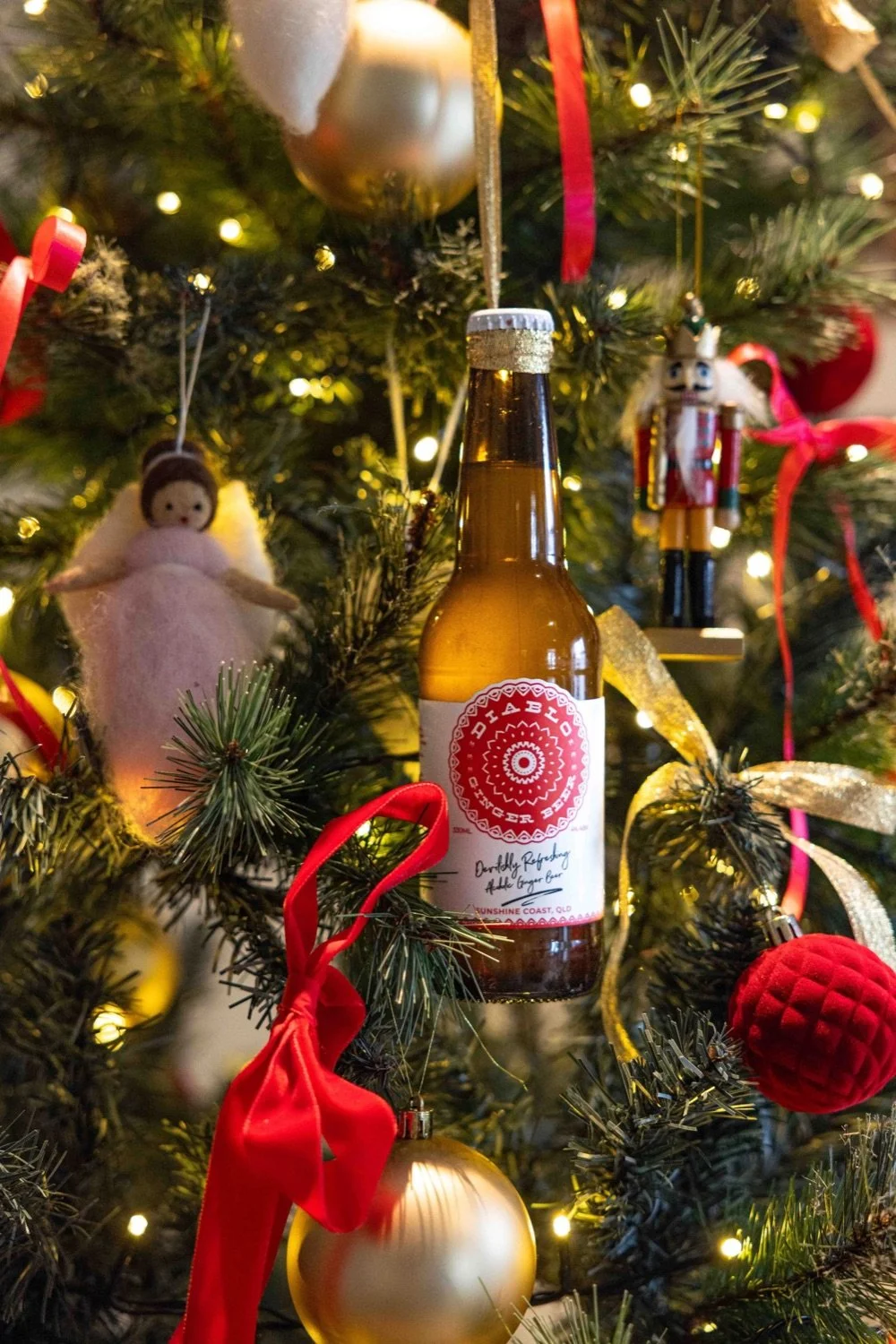 A Christmas tree decorated with ornaments such as gold and red balls, ribbons, and figurines. A bottle of ginger beer hangs on the tree among the decorations.