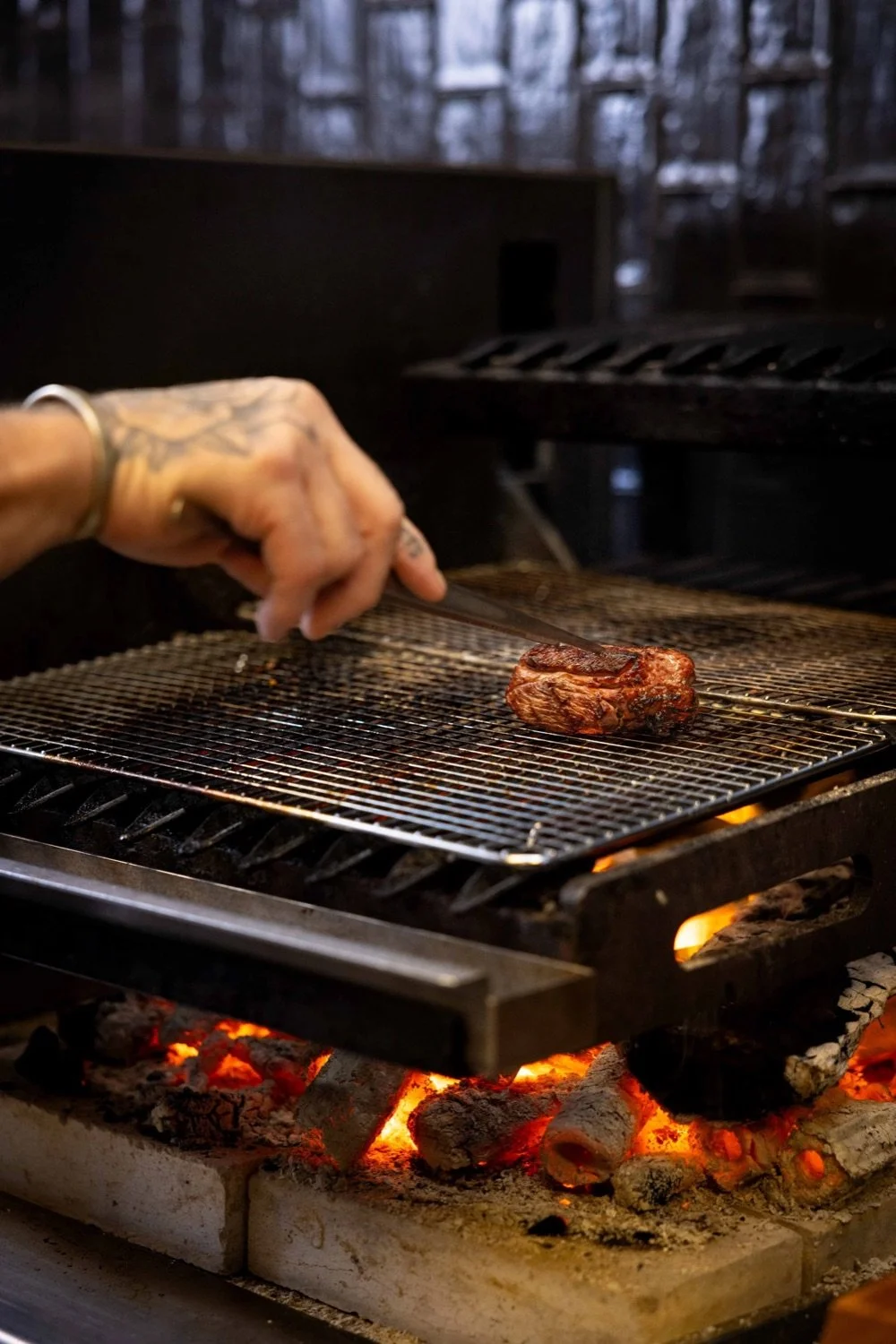 A person with a tattooed hand is using tongs to handle a seared piece of steak on a grill rack above charcoal embers in a barbecue smoker.