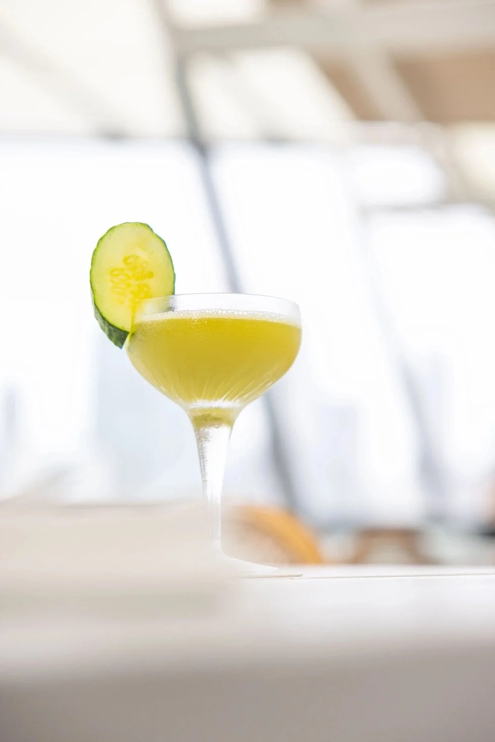 A cocktail in a coupe glass garnished with a cucumber slice on the rim, with a blurred bright indoor background.