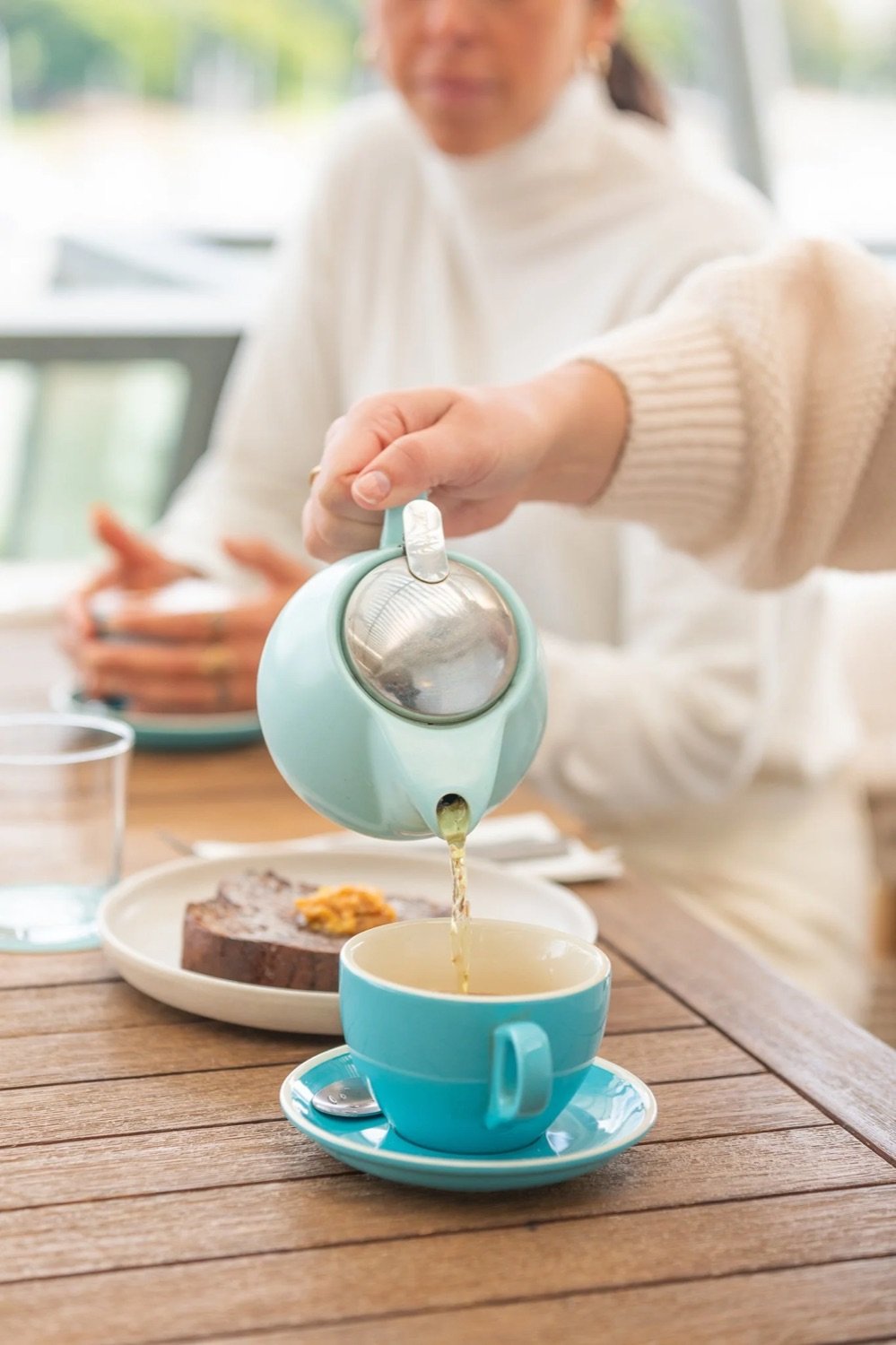 Person pouring tea from a teapot into a blue cup on a wooden table with a plate of toast and a glass of water.