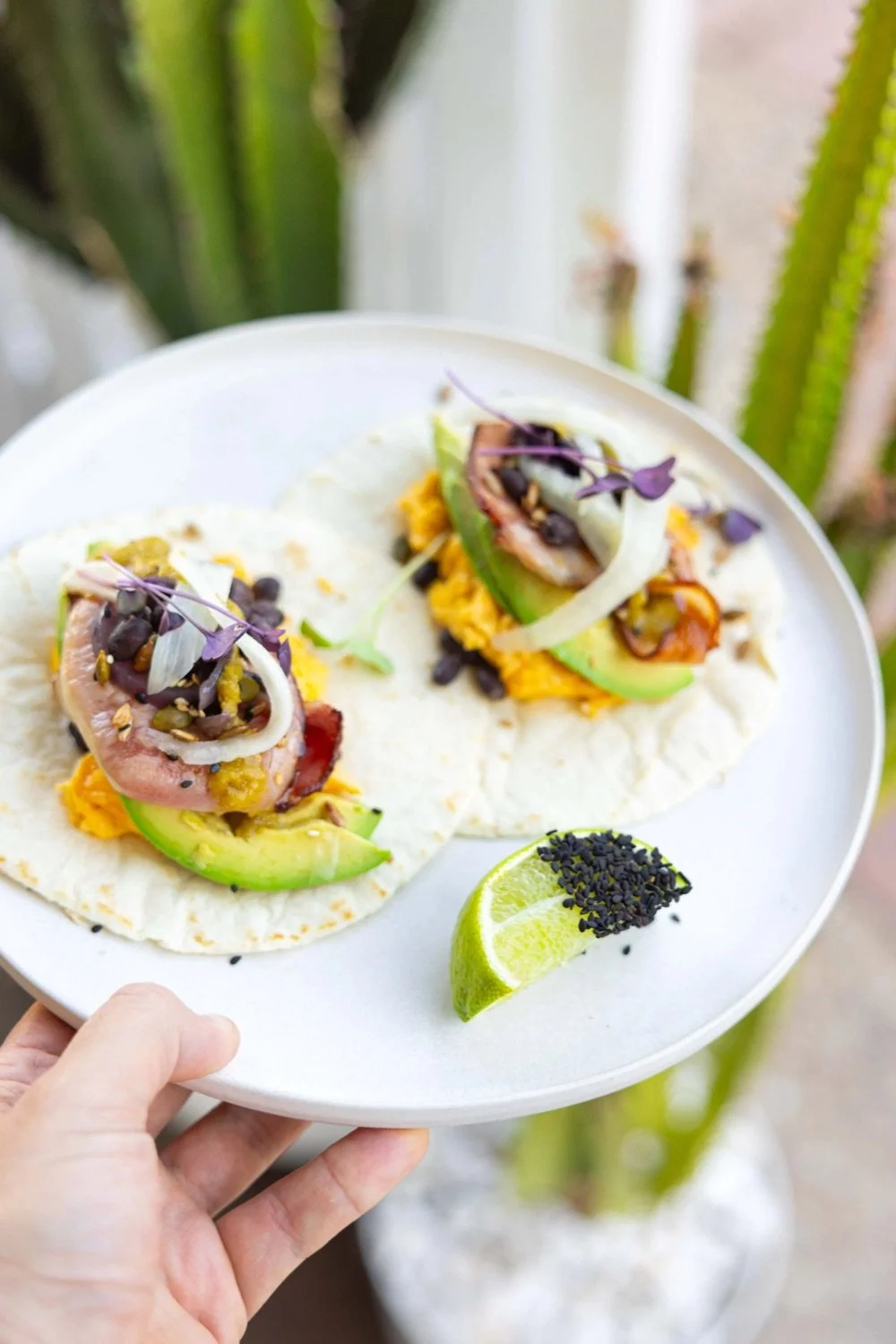 Two tacos with avocado, onions, black beans, shredded cheese, and microgreens on a white plate, with a lime wedge topped with black sesame seeds.