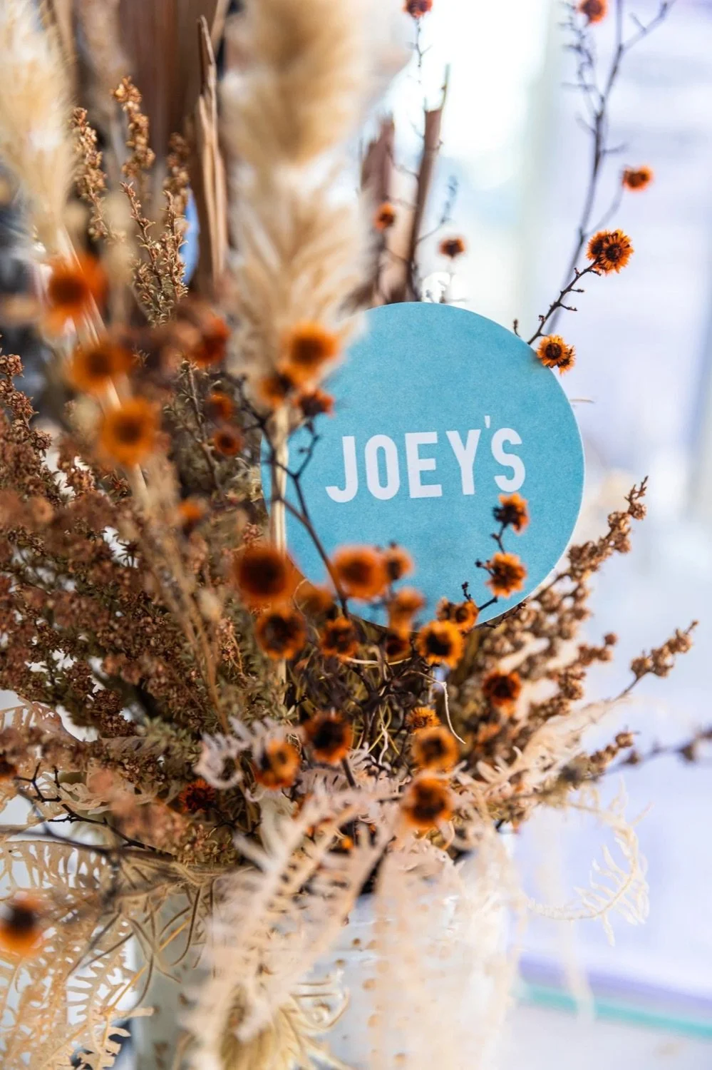 A flower arrangement with a blue circular tag that reads 'Joey's', surrounded by dried flowers and grasses in shades of orange, brown, and beige.
