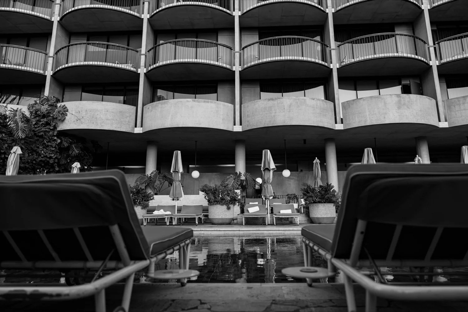 Black and white photo of a hotel or apartment building with multiple curved balconies, outdoor lounge chairs, umbrellas, and potted plants near a pool.