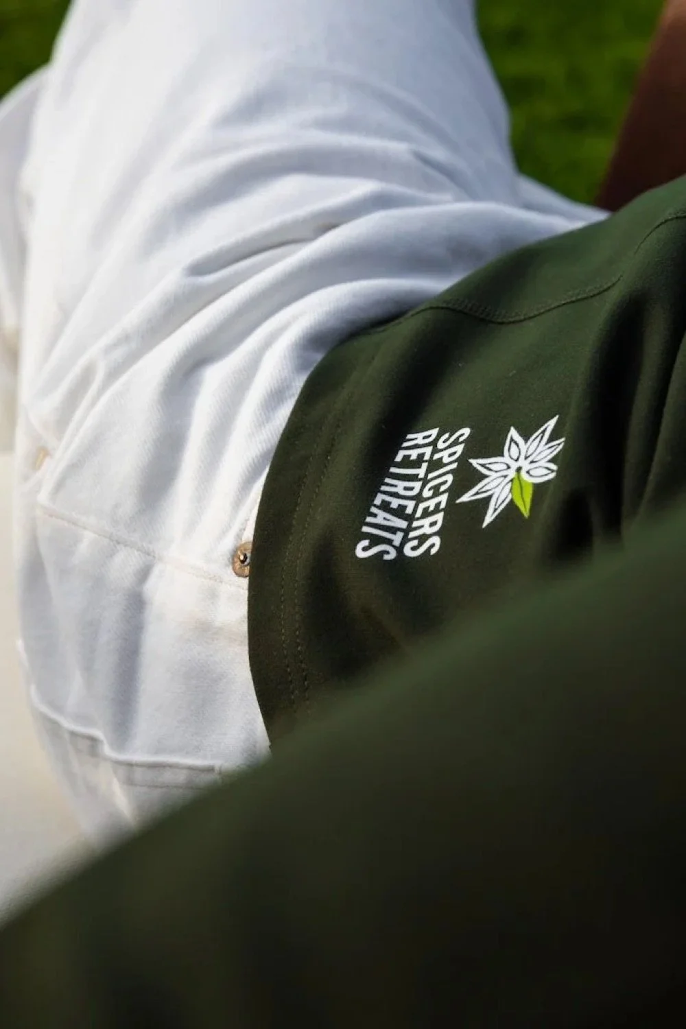 Close-up of a person wearing a dark green jacket with the Sprayberries Retreat logo on the sleeve and white pants, outdoors on a grassy area.