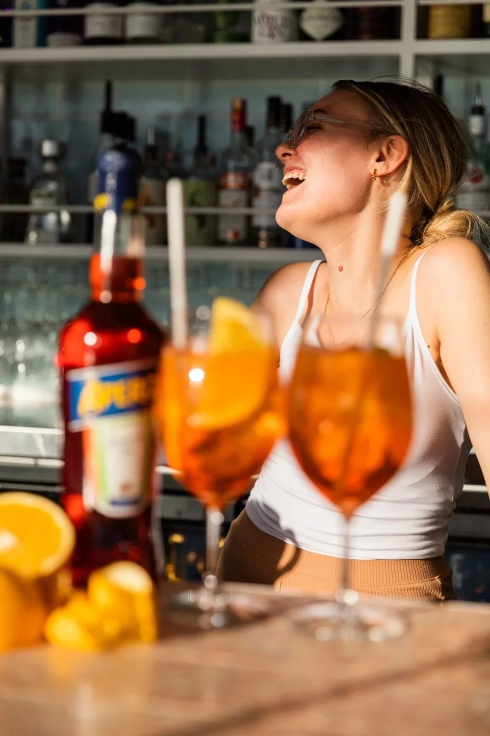 A woman laughing in a bar or kitchen, with bottles and glasses of orange-colored drinks and lemons in the foreground.