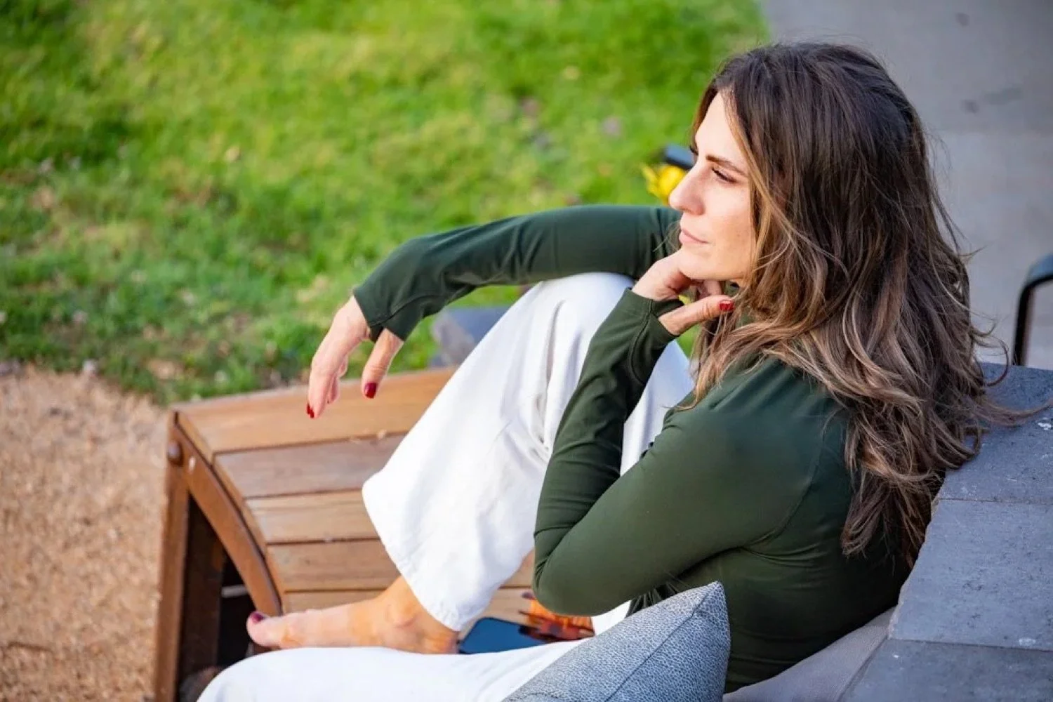 Woman sitting on a wooden bench outdoors with greenery in the background, wearing a green jacket and white pants, resting her chin on her hand and looking thoughtfully to the side.