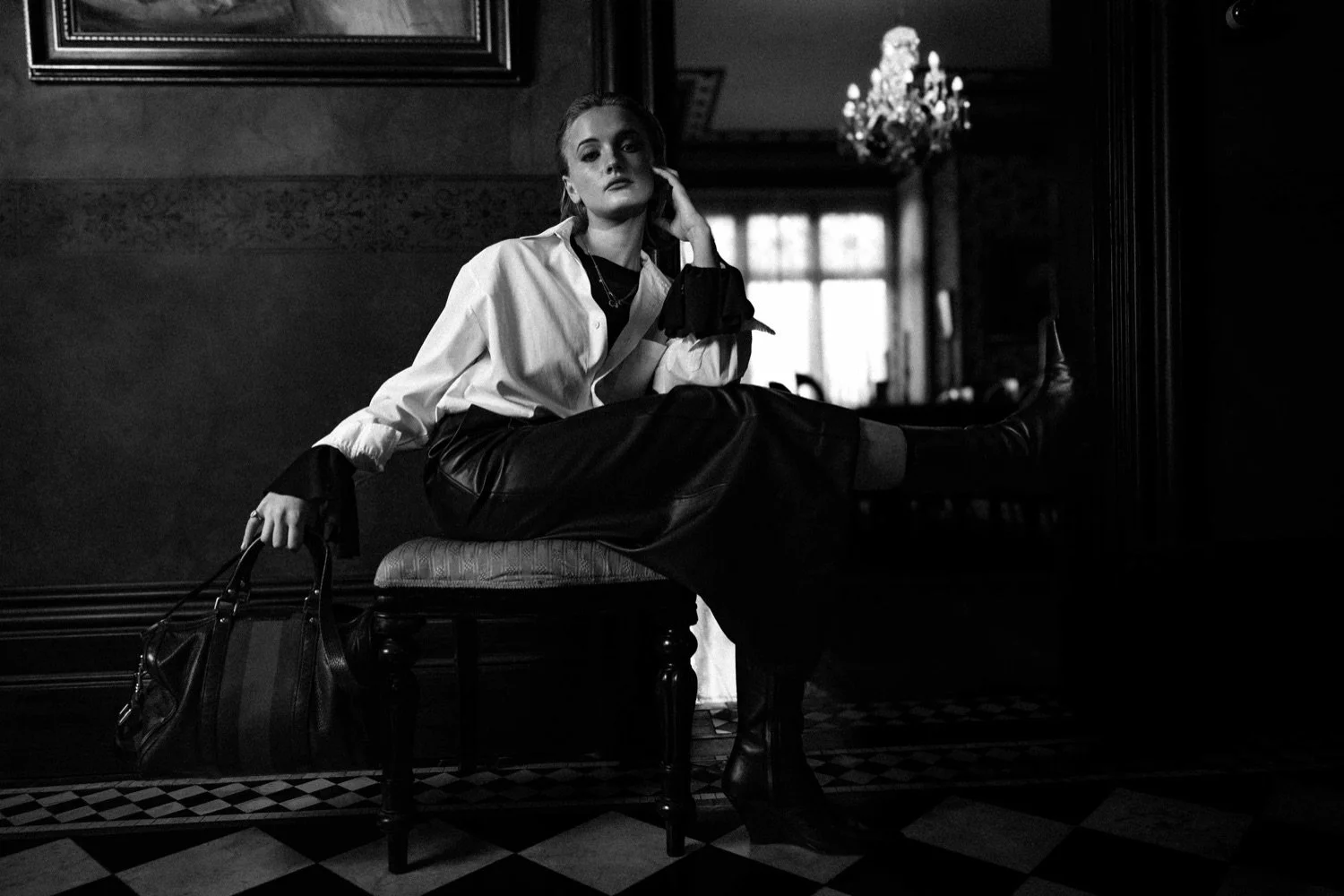 A woman in a white blouse and dark leather pants sits on a cushioned bench, holding a handbag in a dimly lit room with ornate décor and a chandelier.