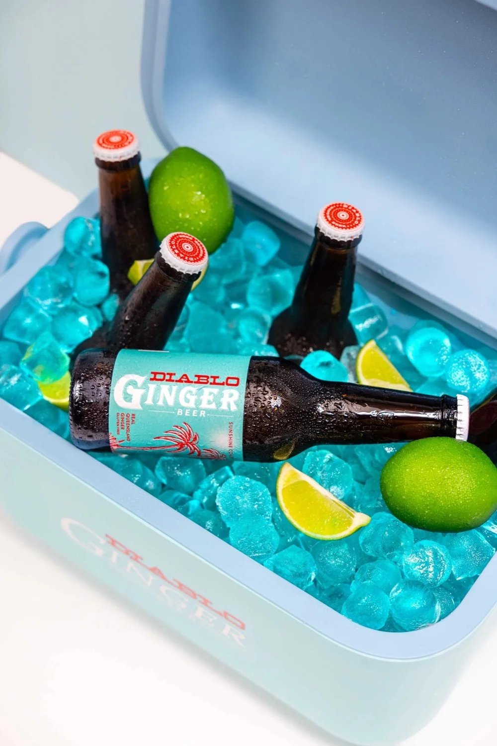 A light blue cooler filled with ice, three bottles of Diablo Ginger Beer, two limes, and a green lime-shaped object.