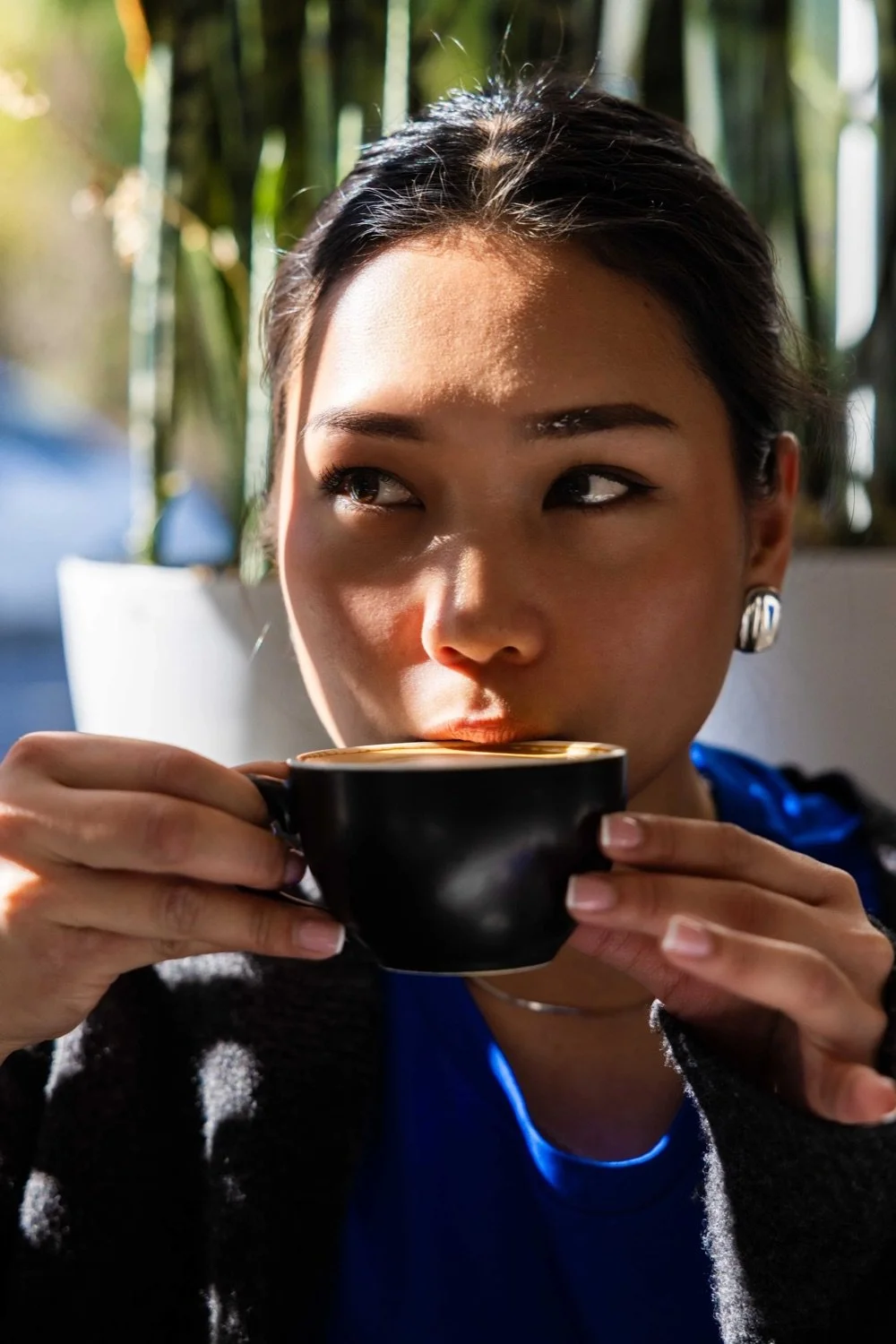 A woman with dark hair and earrings is drinking coffee from a black cup, illuminated by sunlight, with green plants in the background.