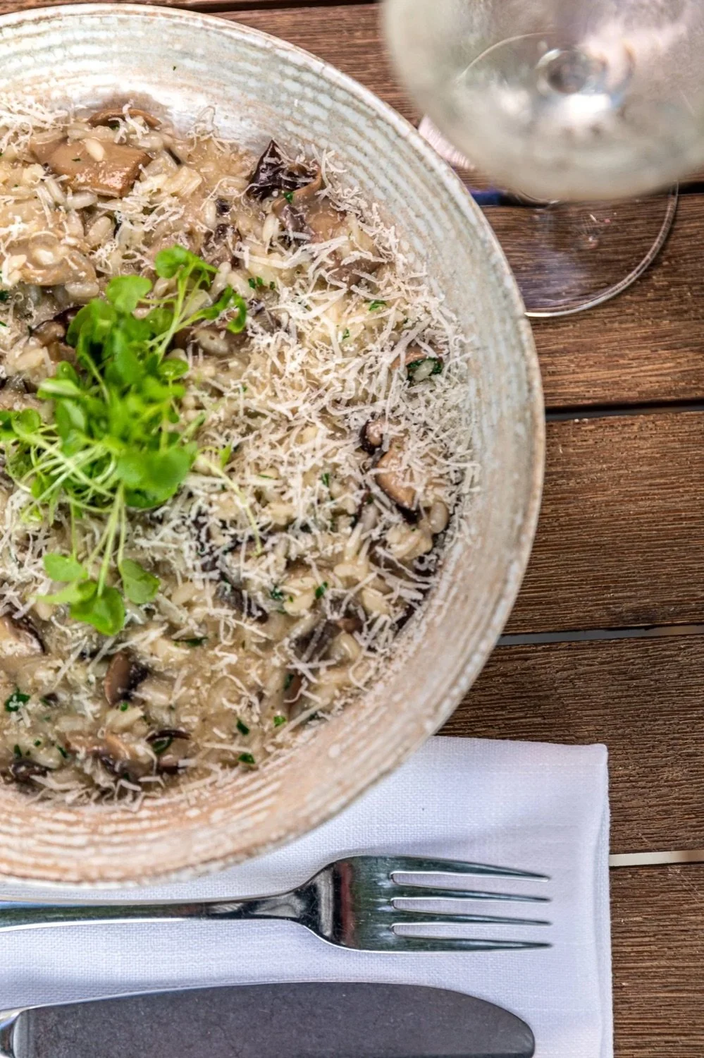 Creamy mushroom risotto garnished with fresh herbs and grated cheese on a clear glass bowl, with a fork and napkin on a wooden table and a glass of white wine.