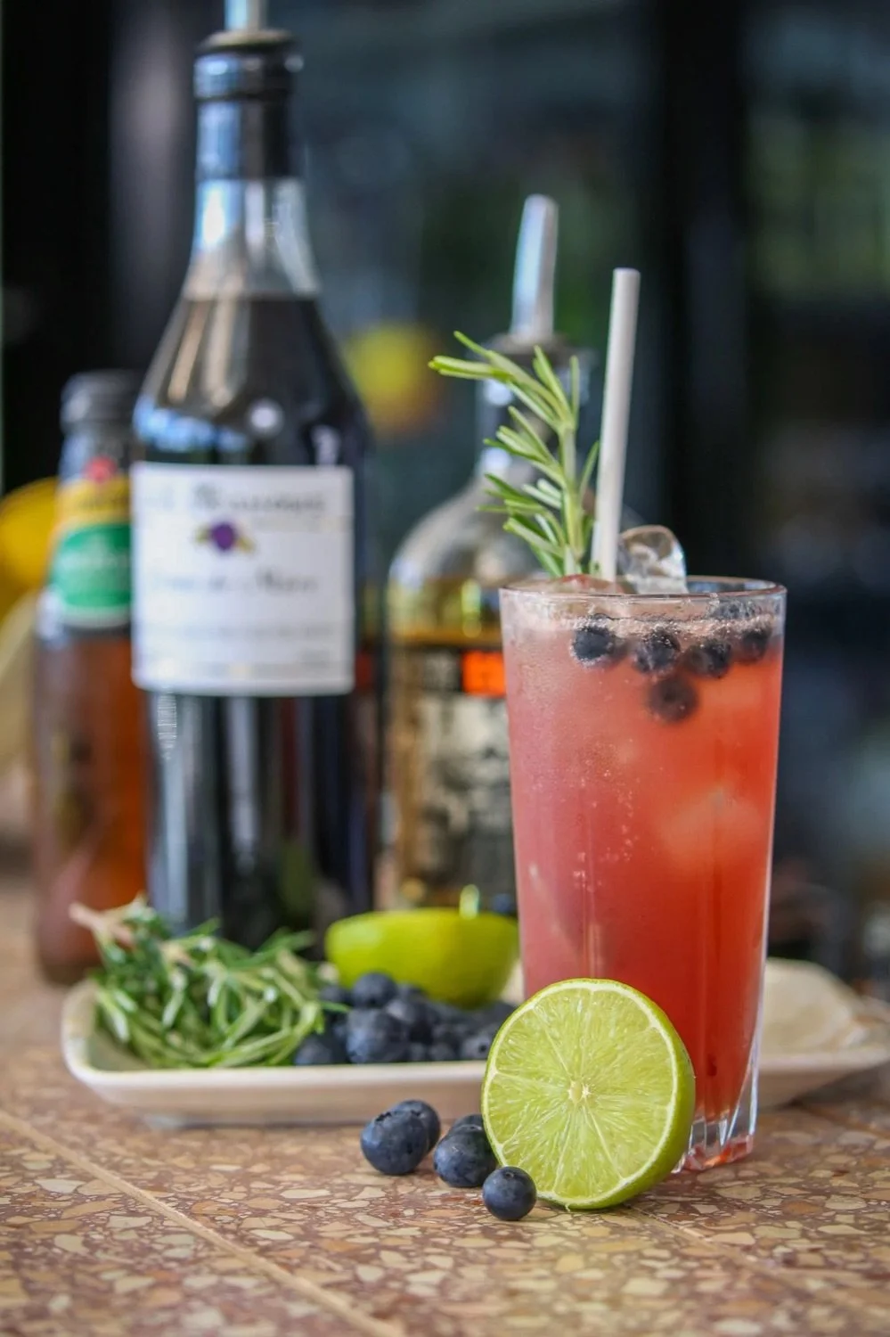 A tall glass of pink berry cocktail garnished with a sprig of rosemary and blueberries, with a fresh lime half and blueberries on a patterned surface, and bottles of liquids in the background.