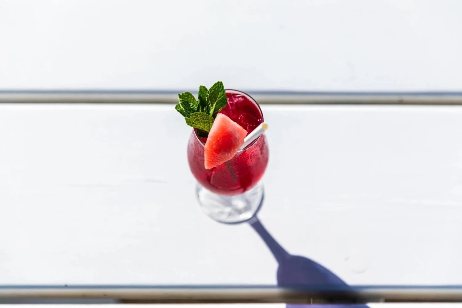 Red cocktail garnished with mint leaves, watermelon wedge, and a straw, casting a shadow on a white surface.