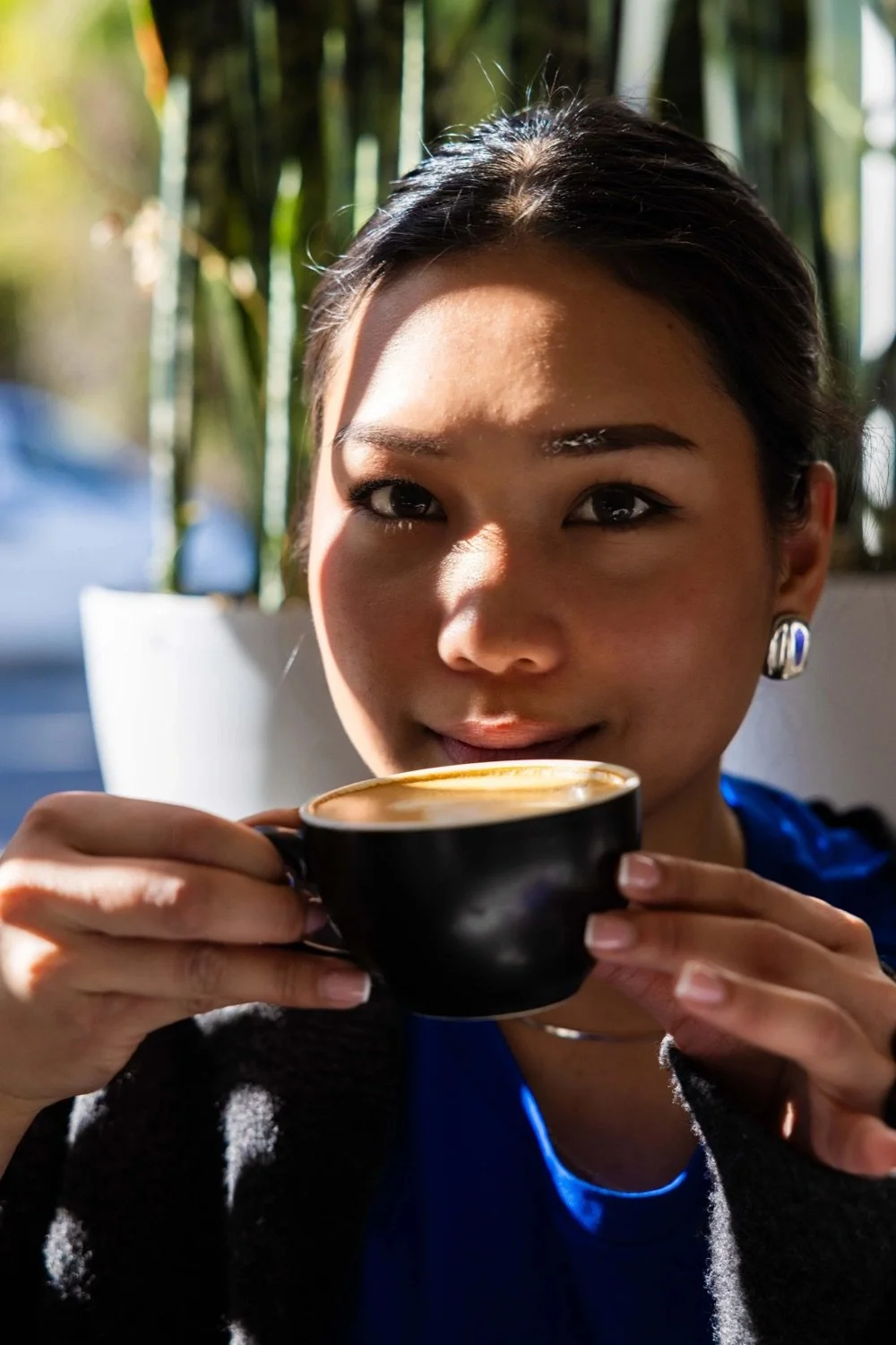 A woman with dark hair and earrings holding a black cup of coffee or latte, smiling softly, with sunlight illuminating her face and a background of green plants.