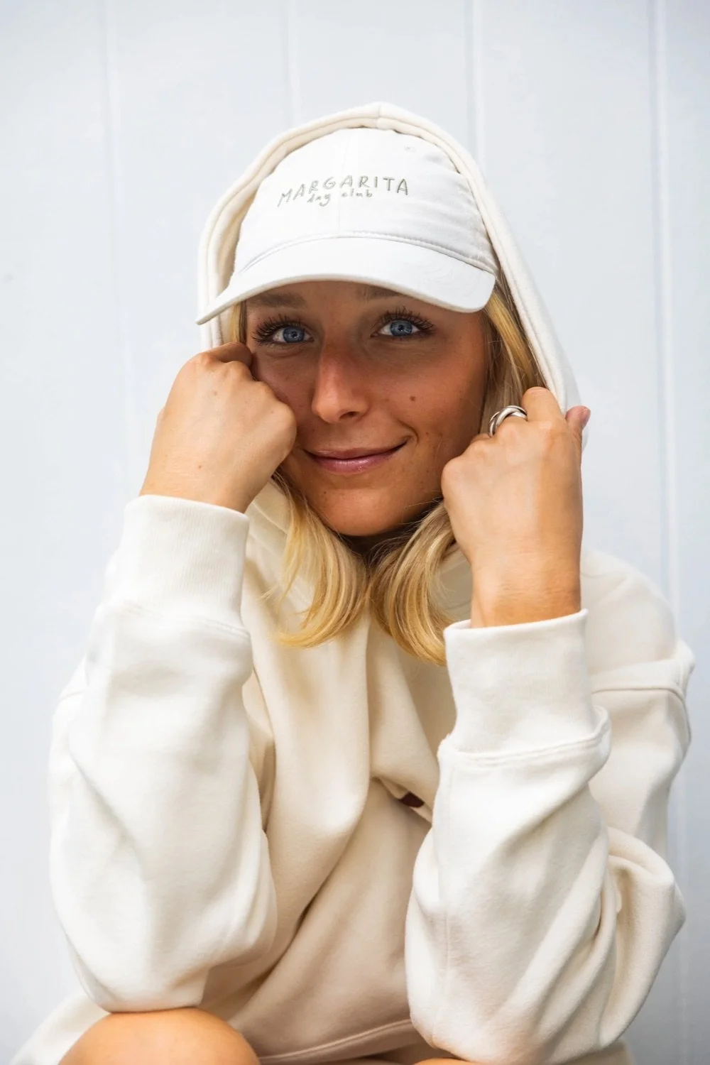 A woman with blonde hair and blue eyes wearing a white hoodie and a white cap that says 'MARGARITA' on it, smiling warmly at the camera, with her fists gently resting on her cheeks.