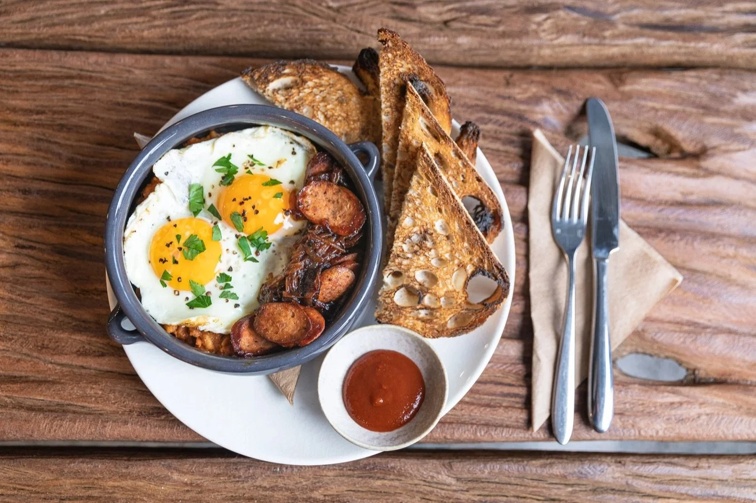 A plate of breakfast with two fried eggs topped with herbs, crispy bacon, roasted potatoes, toasted bread slices, and a small bowl of red sauce on a wooden table.