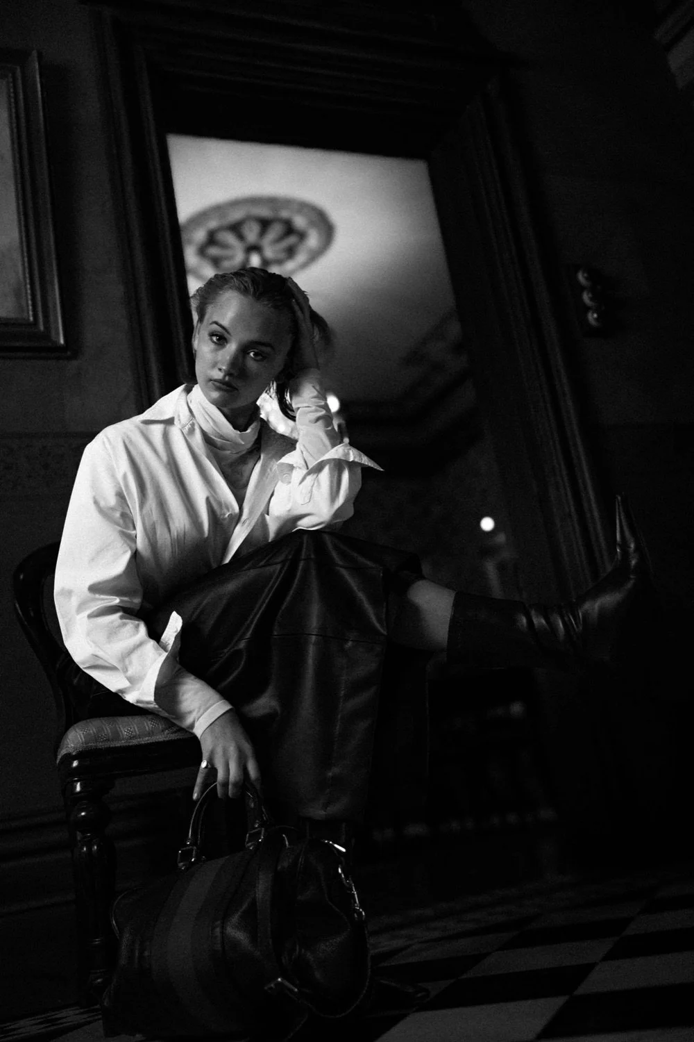 A black and white photo of a woman sitting on an ornate chair in a dimly lit room, with a mirror reflecting her face and part of the ceiling.