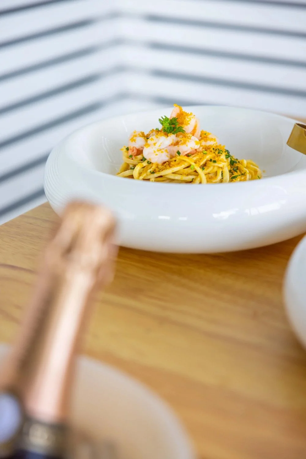 A white bowl of shrimp pasta garnished with herbs and lemon zest, sitting on a wooden table.
