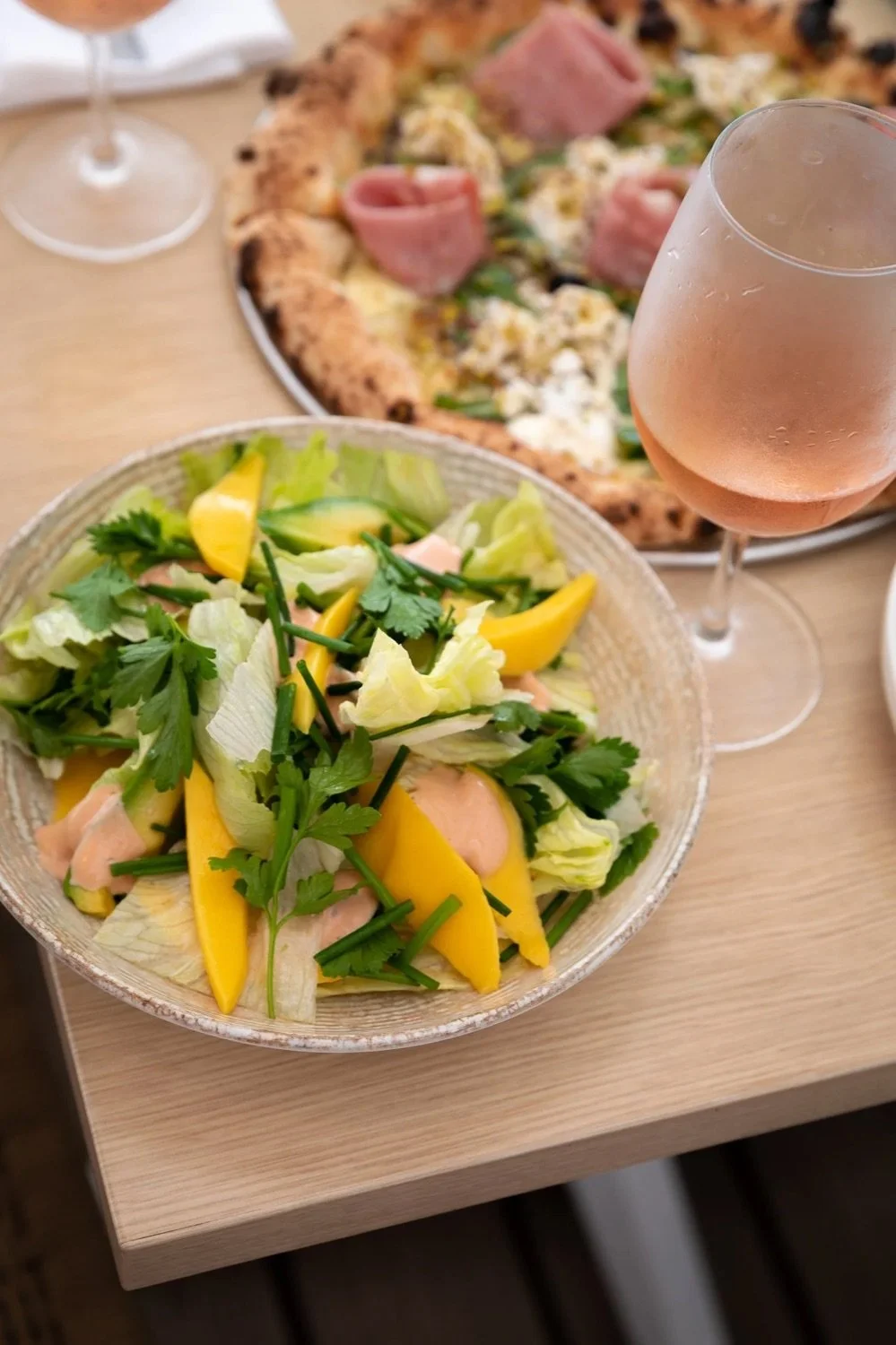 A bowl of mixed salad with lettuce, yellow bell peppers, cilantro, and a creamy dressing on a wooden table. In the background, there is a pizza with toppings that appear to include ham, cheese, and possibly greens, alongside a glass of rosé wine.