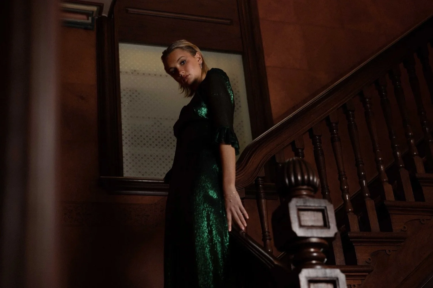 Woman in an elegant black dress with green accents stands on a wooden staircase inside a dimly lit interior.