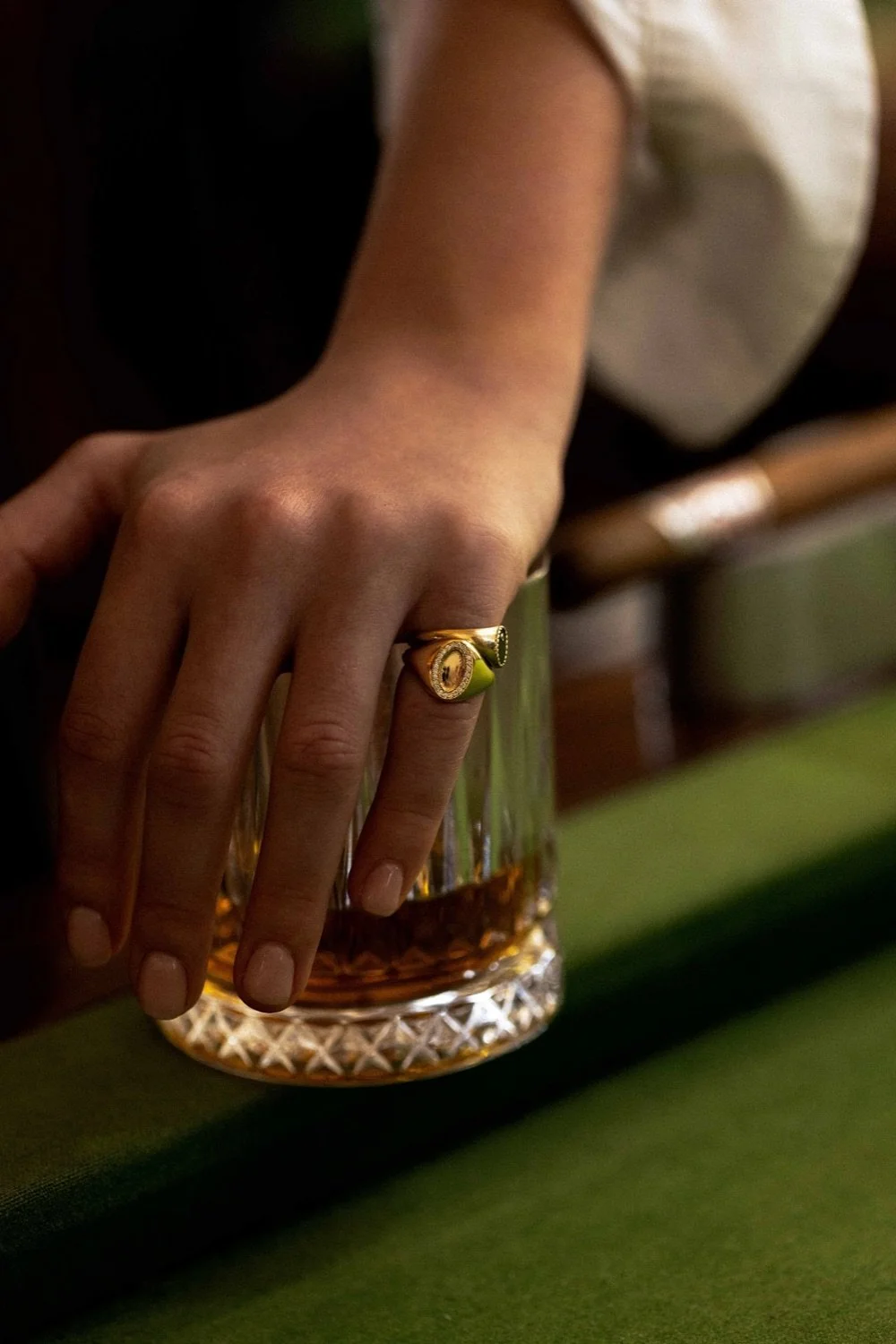 Close-up of a person's hand with a gold ring, resting on the rim of a glass of whiskey on a green surface.