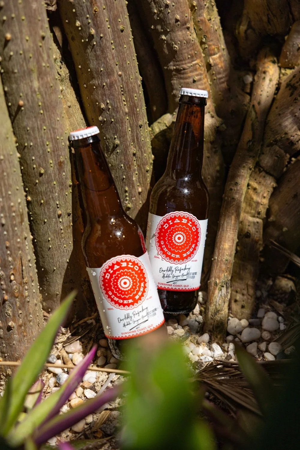 Two brown glass beer bottles with white and red labels, resting against a background of stacked firewood and some greenery in the foreground.