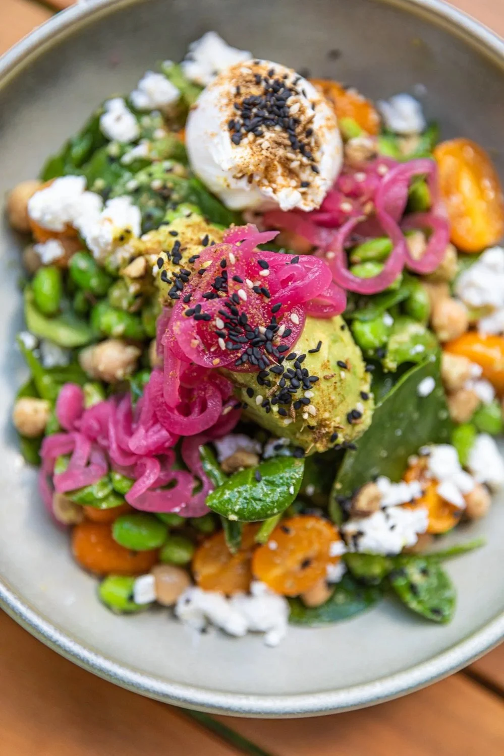 Close-up of a colorful salad with avocado, pickled onions, cherry tomatoes, chickpeas, fresh greens, crumbled cheese, and a poached egg topped with black sesame seeds in a bowl.