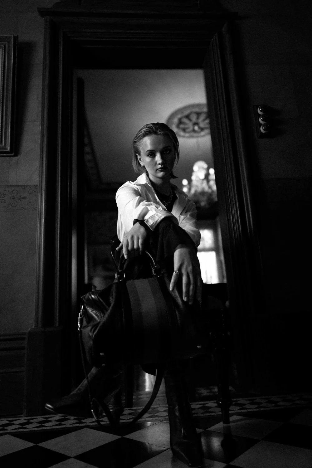 A black-and-white photo of a young woman sitting on a chair in front of a mirror, holding a handbag, wearing boots, with a serious expression, in a dimly lit room.