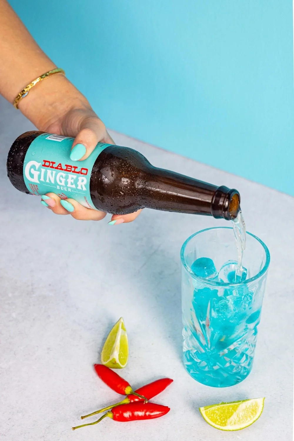 Person pouring Diablo Ginger Beer into a glass with ice, accompanied by lime wedges and red chili peppers on a light gray surface, with a blue wall background.