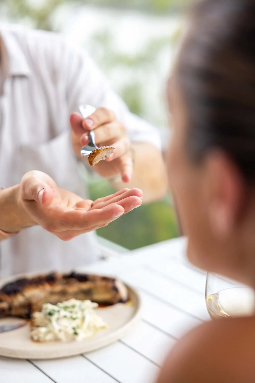 Person handing a piece of cooked meat on a fork to another person at a dining table with food in the background.