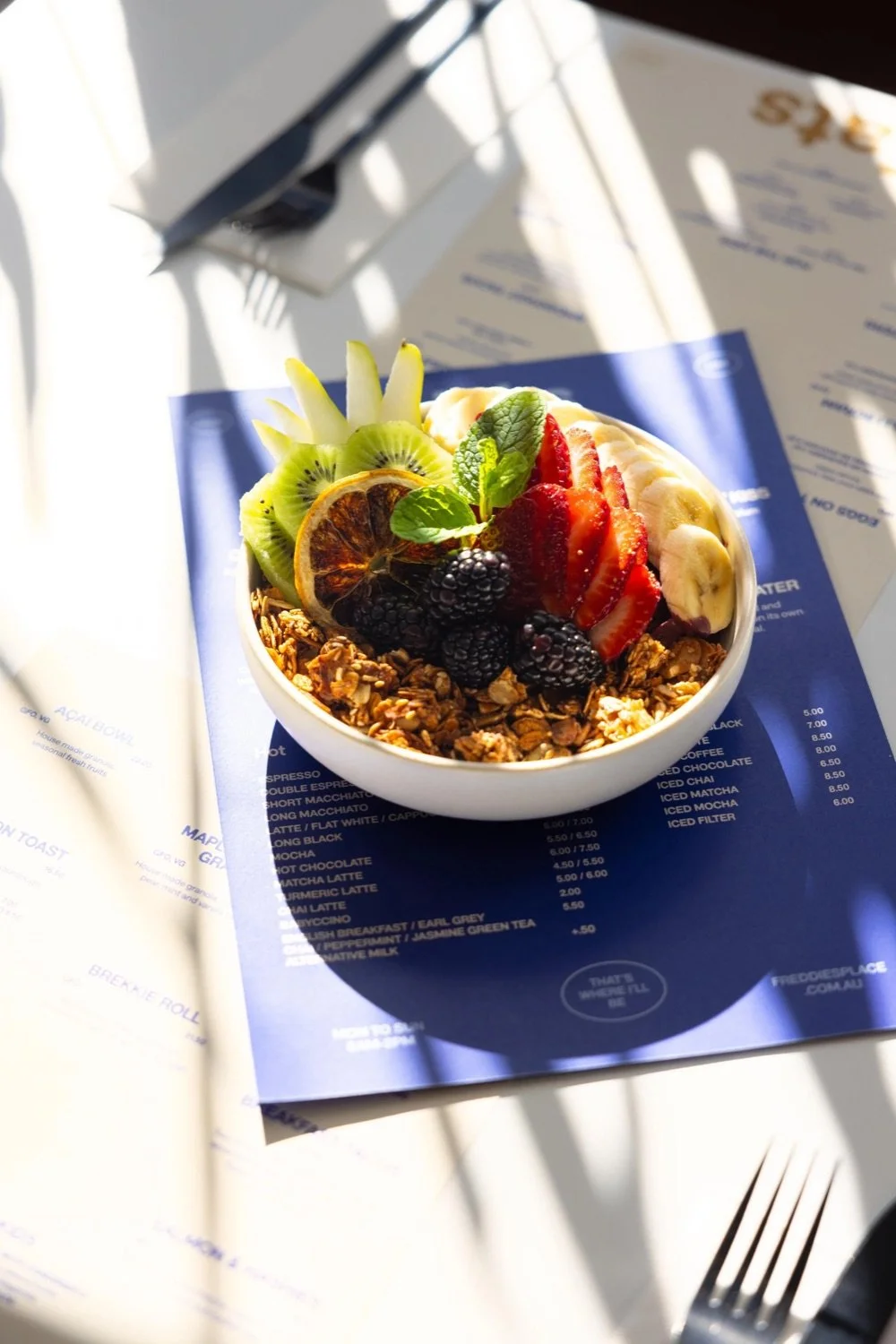 Bowl of fresh fruit topped with sliced strawberries, blueberries, blackberries, kiwi, and a dried lemon slice, served over granola and garnished with mint leaves, placed on a blue menu at a dining table with cutlery.