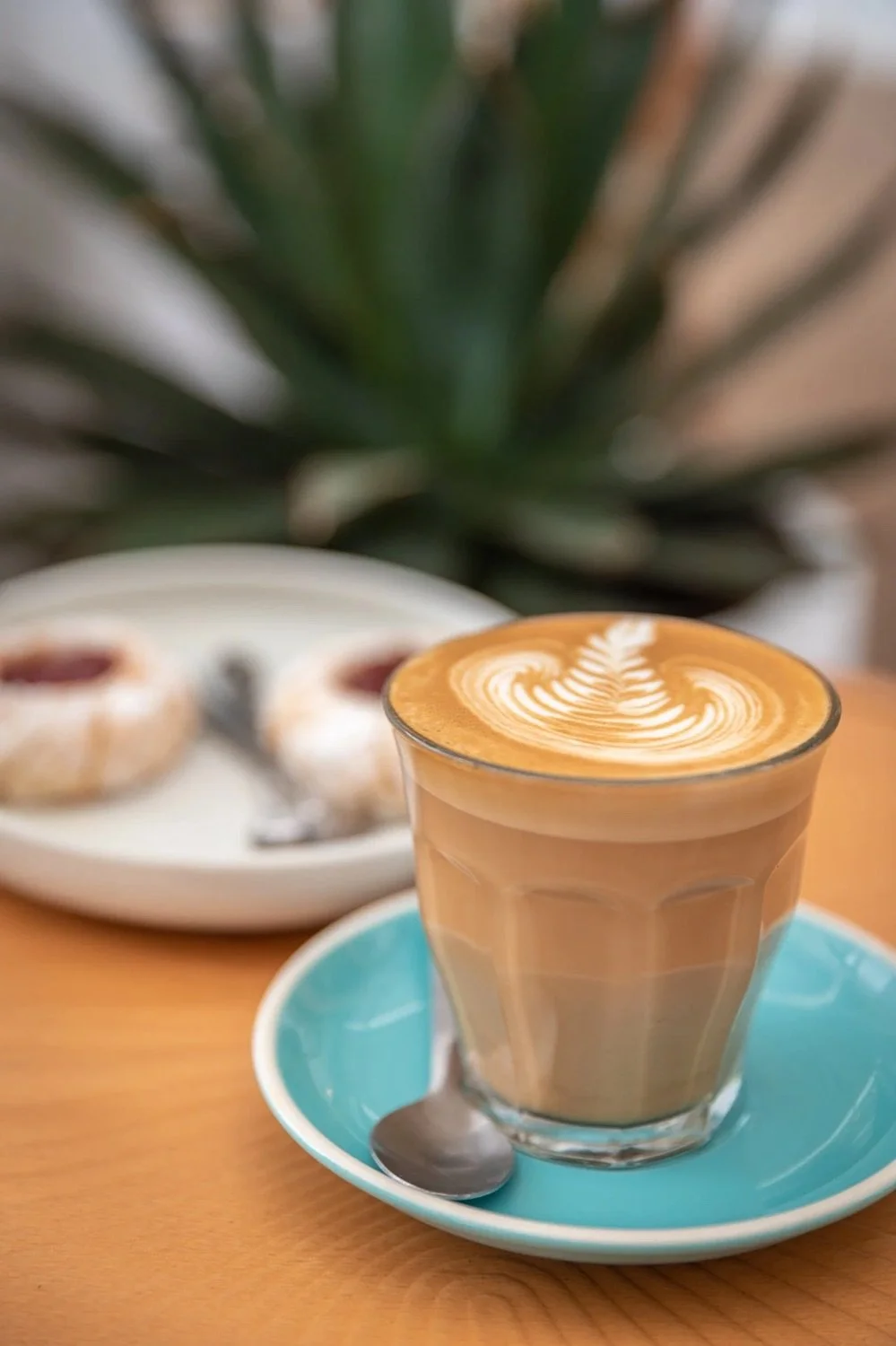 A glass of caramel-colored latte with latte art on top, a blue saucer, and a small spoon, with a plate of donuts and a pineapple in the background.