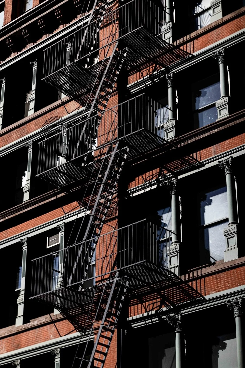 Multiple fire escapes on a brick building with large windows, casting shadows on the facade.