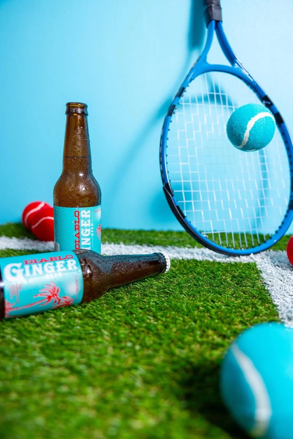 Two bottles of Diablo Ginger beer, tennis rackets, and tennis balls on artificial turf against a light blue wall.