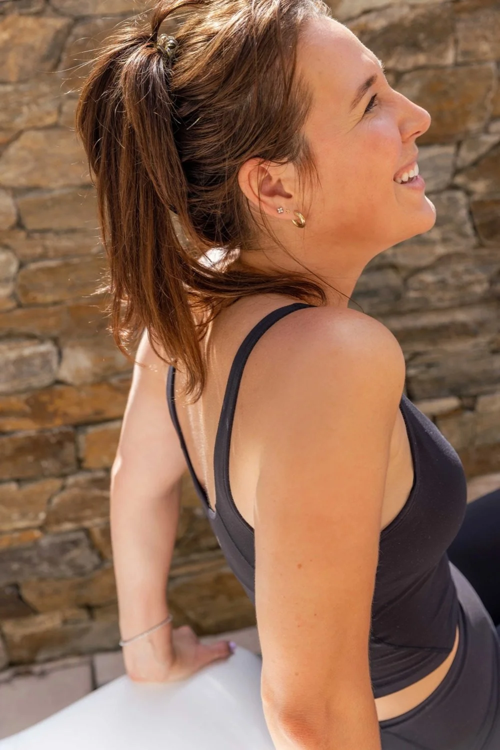 A woman with brown hair tied in a ponytail, smiling with her eyes closed, wearing a black tank top and earrings, against a stone wall background.