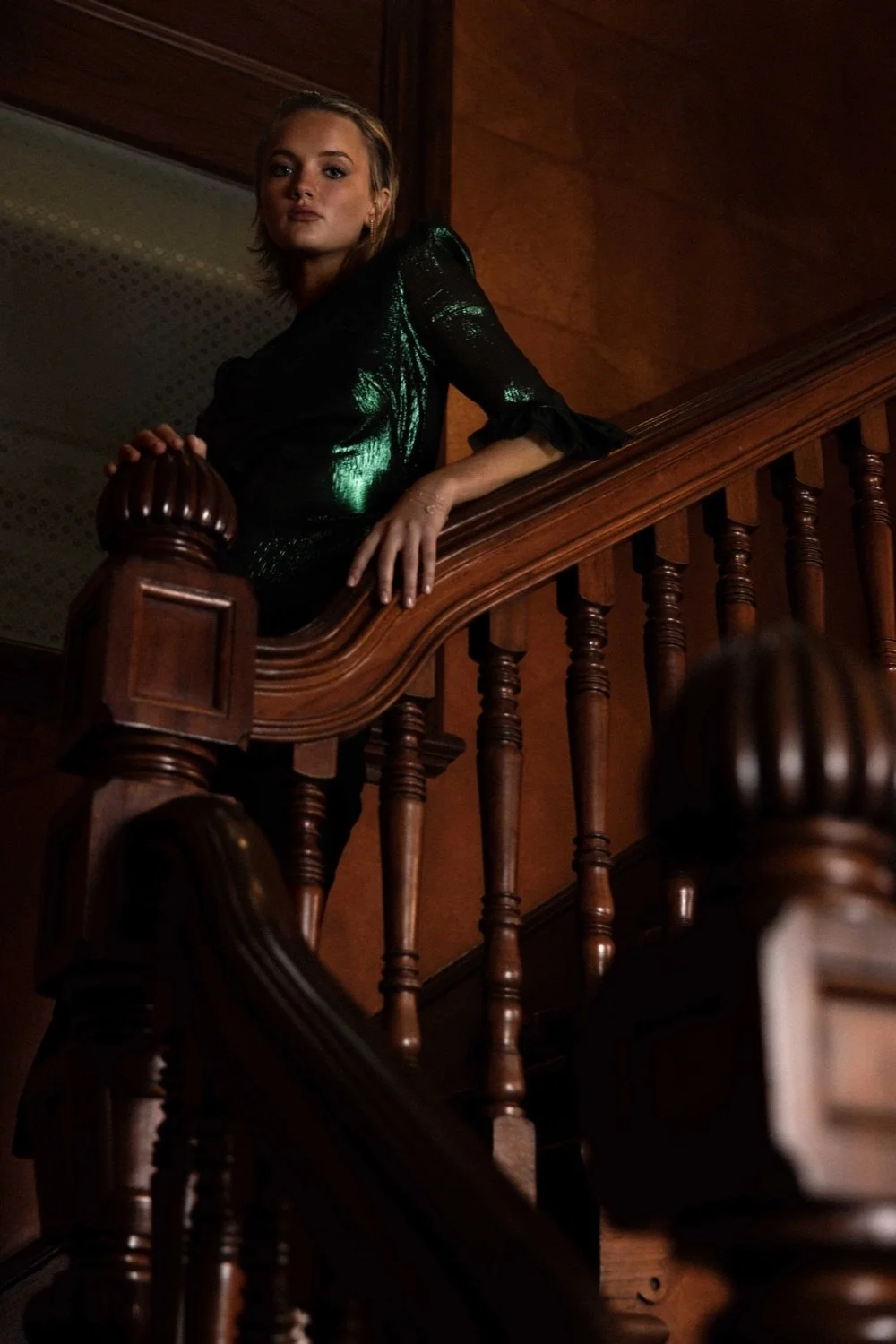 A woman with light-colored hair in a black shiny dress stands on a wooden staircase, looking at the camera. The staircase has carved wooden railings and the background is warm-toned wood paneling.