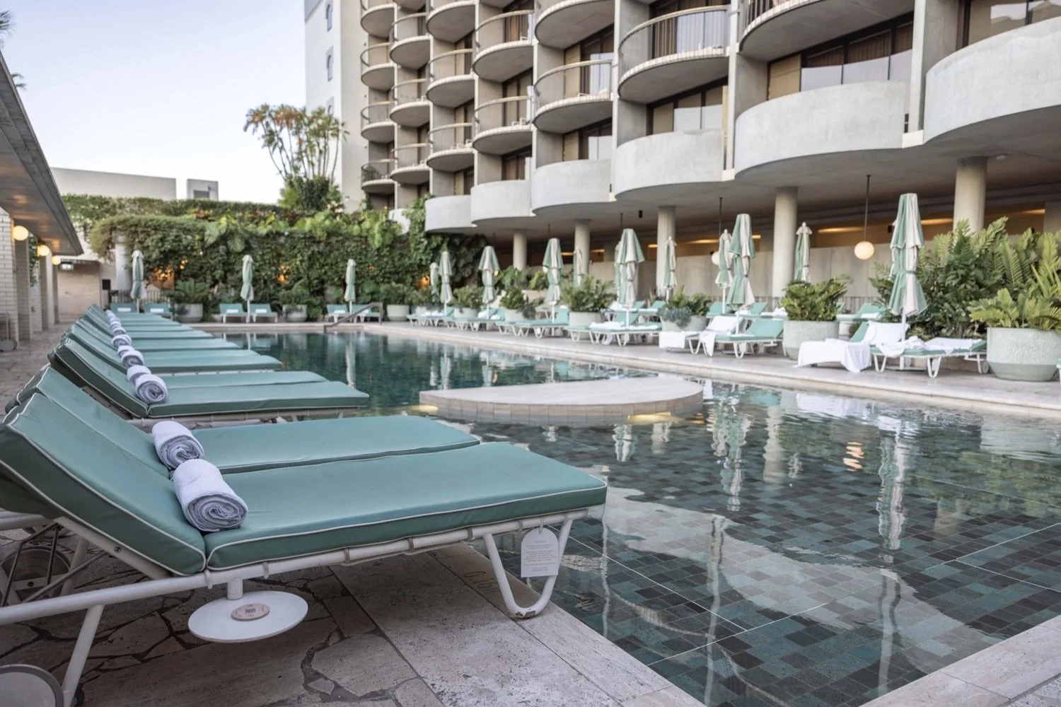 Empty outdoor swimming pool area with lounge chairs, umbrellas, and towels on a hotel patio, with a modern building in the background.