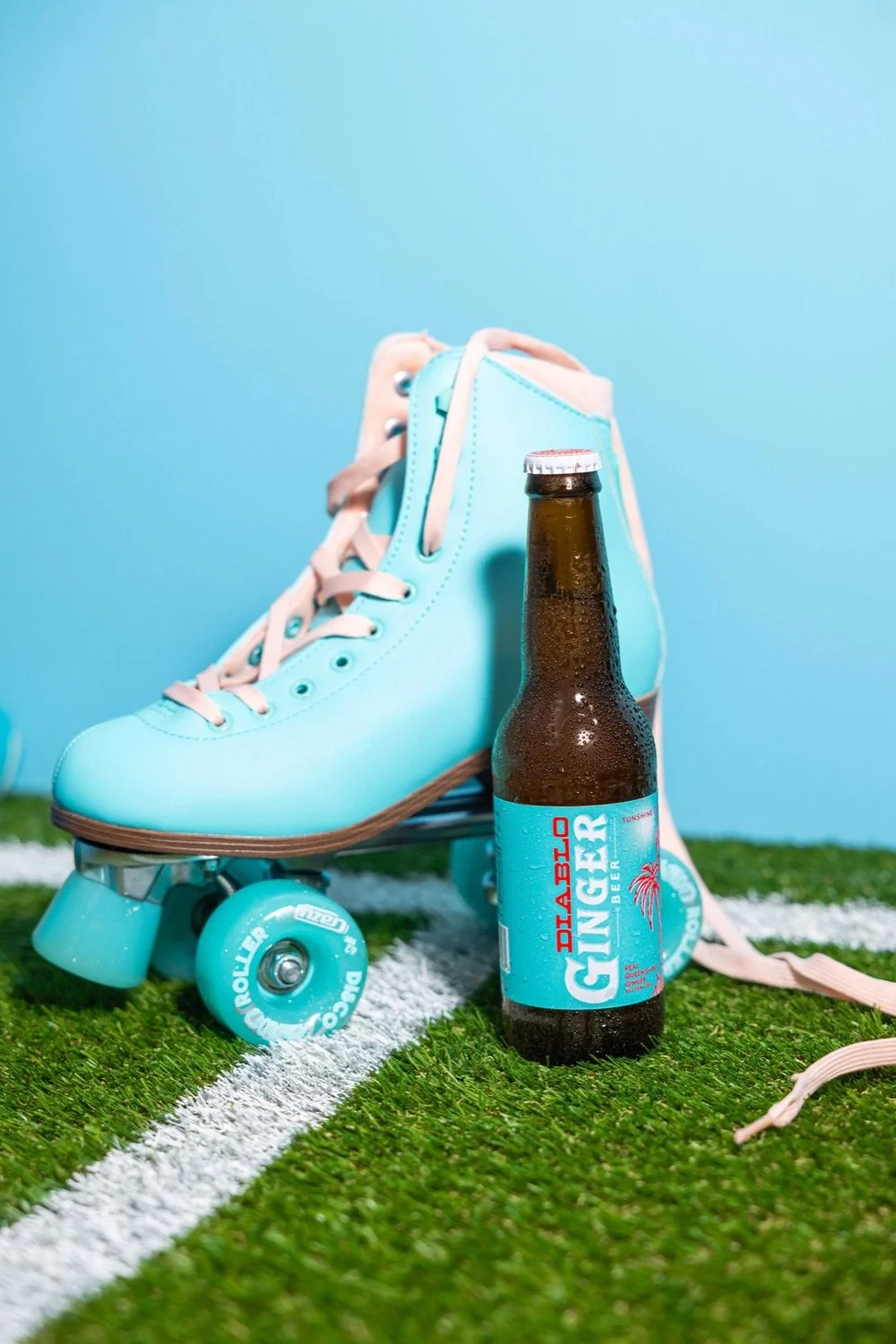 A mint green roller skate, a bottle of DABLO GINGER beer, and a pink lace-up shoe on green turf with white lines and a light blue background.