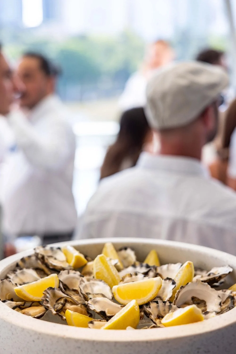 A plate of raw oysters garnished with lemon wedges in the foreground, with blurred people in white chef uniforms and a chef's hat engaging in conversation in the background.