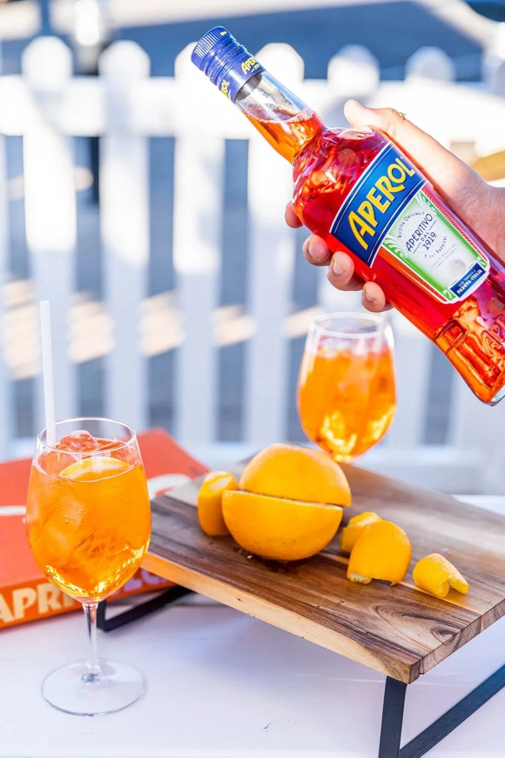 A hand pouring Aperol from a bottle into a glass with orange-colored drink, two glasses of Aperol spritz, a sliced orange on a wooden board with more orange pieces, on a table outdoor setting.