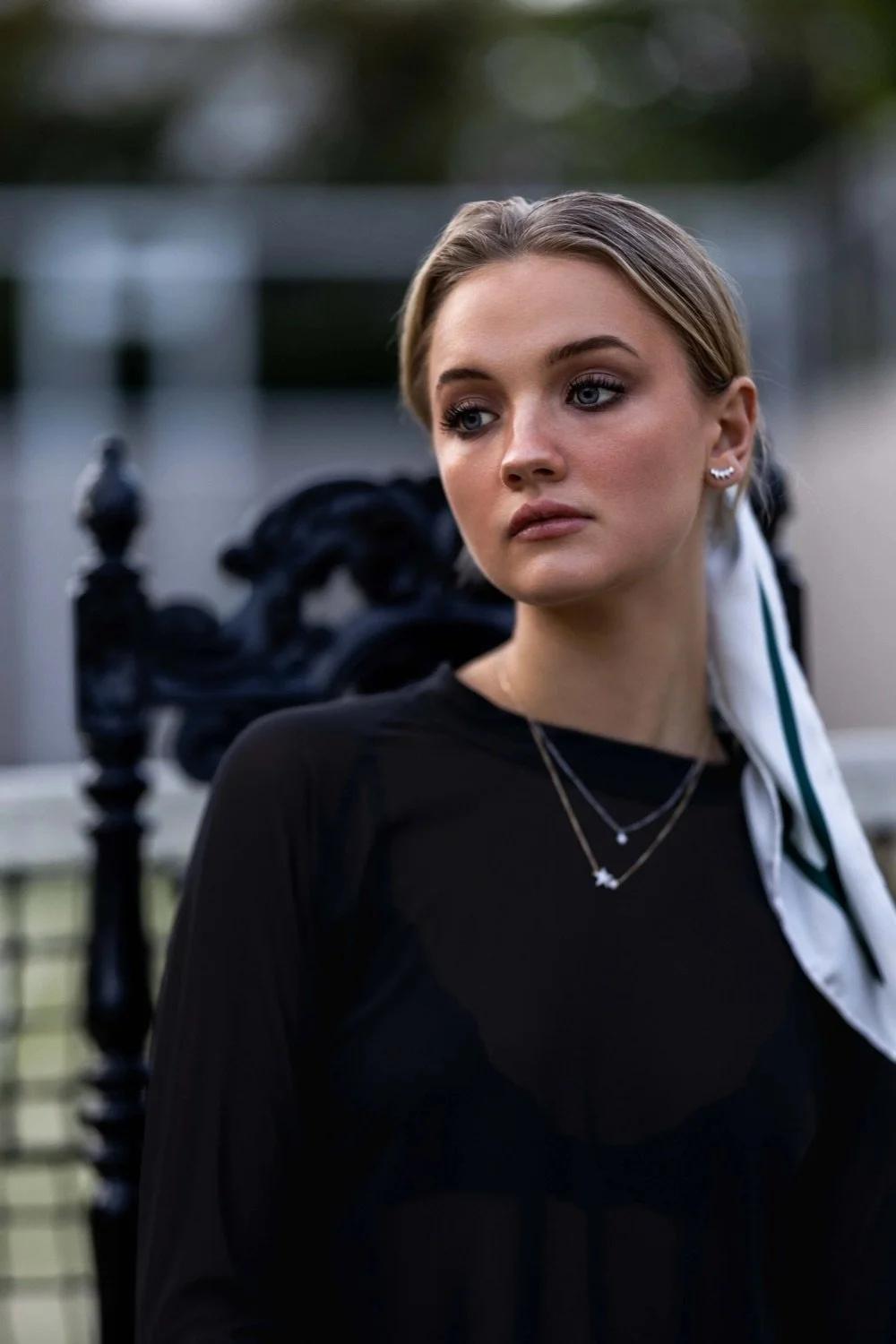 A young woman with blonde hair, wearing earrings and layered necklaces, in a black top with a white scarf tied around her hair, standing outdoors in front of a decorative black wrought iron fence.