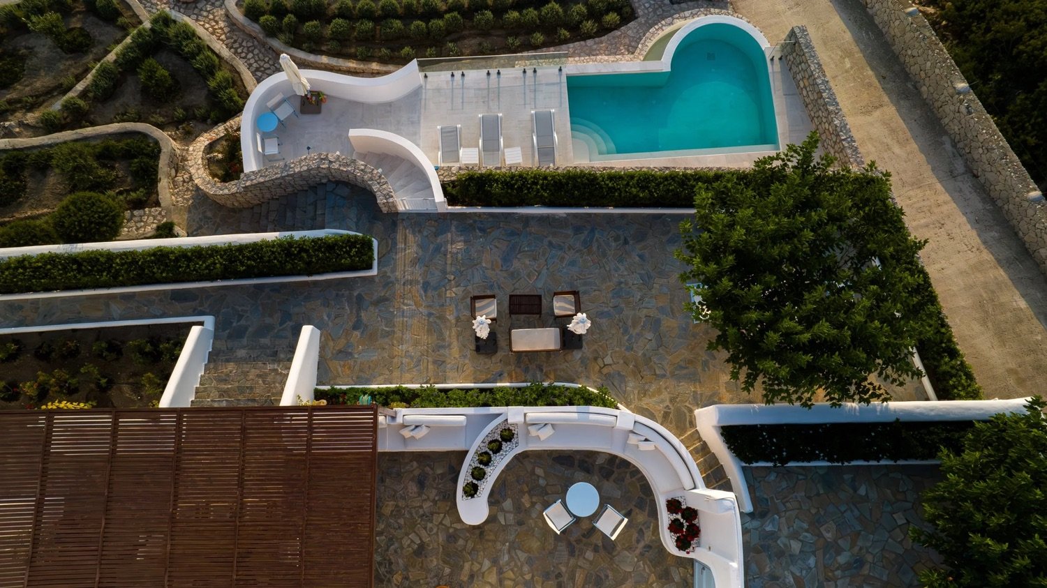 Aerial view of a backyard with a swimming pool, patio, and landscaped garden. The pool is in the top right corner, with a curved edge and steps. The patio features a small table with three chairs and a separate seating area with a round table and two