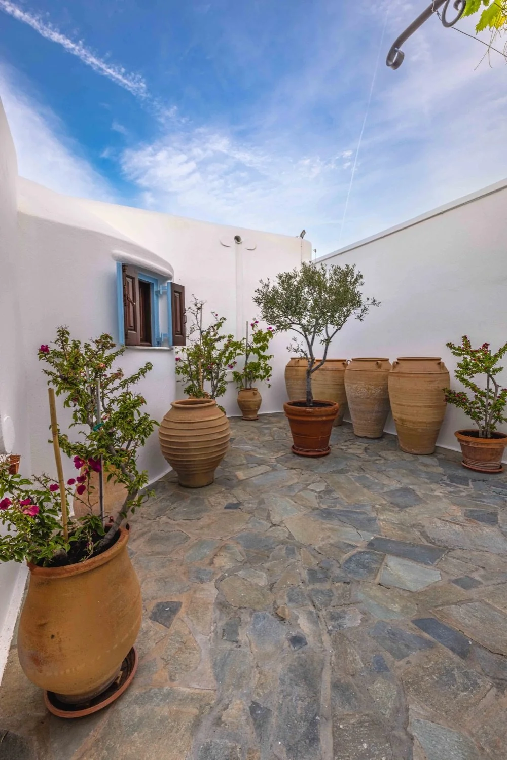 A small outdoor courtyard with white walls, potted plants, and a stone tile floor, under a partly cloudy sky.