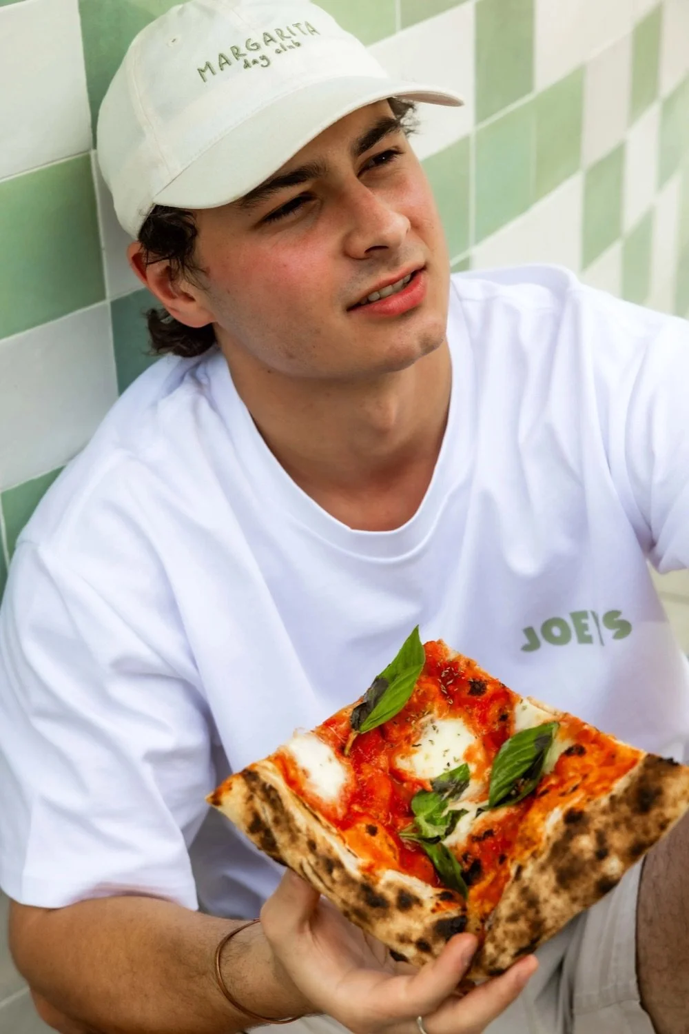 Young man wearing a white cap and white t-shirt holding a large slice of pizza with basil leaves, sitting against green and white tiled wall.