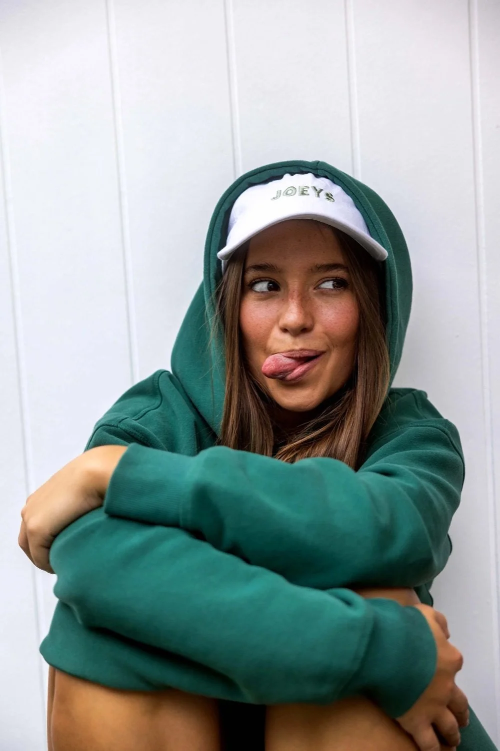 A young woman in a green hoodie and white cap with 'Joey's' embroidered, making a funny face with tongue out and eyes looking sideways, sitting in front of a white wall.