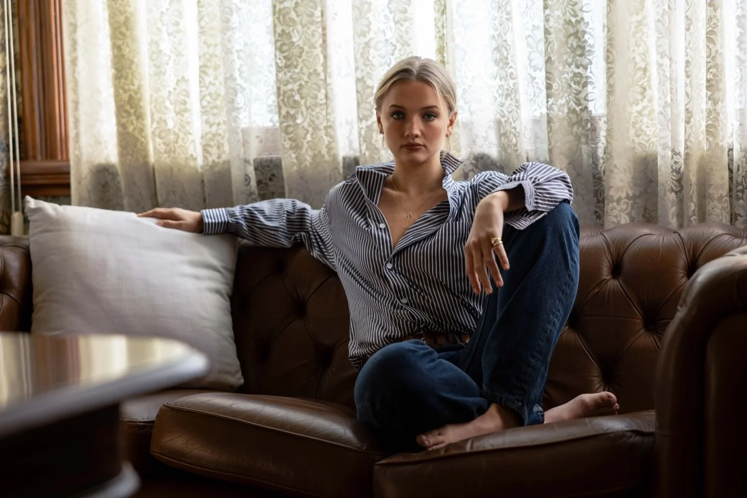 A young woman sitting on a brown leather sofa with one leg crossed over the other, wearing a striped button-up shirt and dark jeans, with a neutral expression, in a room with lace curtains behind her.