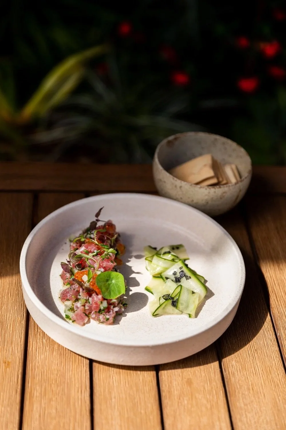 A white round plate with a colorful mixture of chopped raw fish, vegetables, and microgreens on the left, and thinly sliced cucumber with black sesame seeds on the right, placed on a wooden table. In the background, a small bowl with wooden utensils 