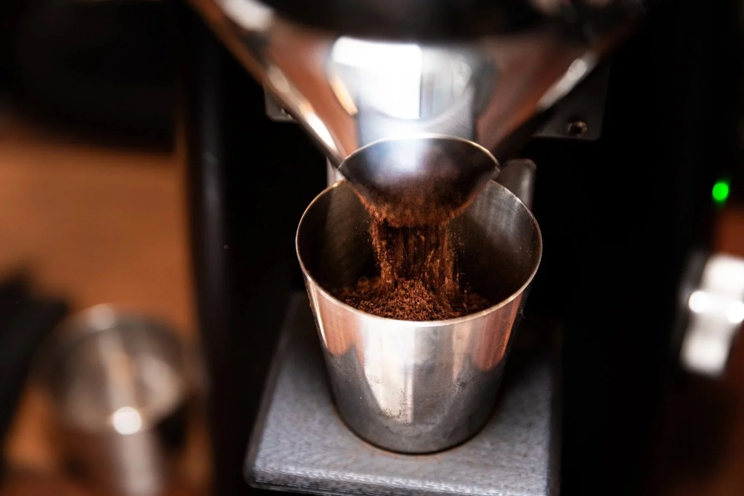Coffee beans are falling from a grinder into a metal container, with a brewing machine visible in the background.