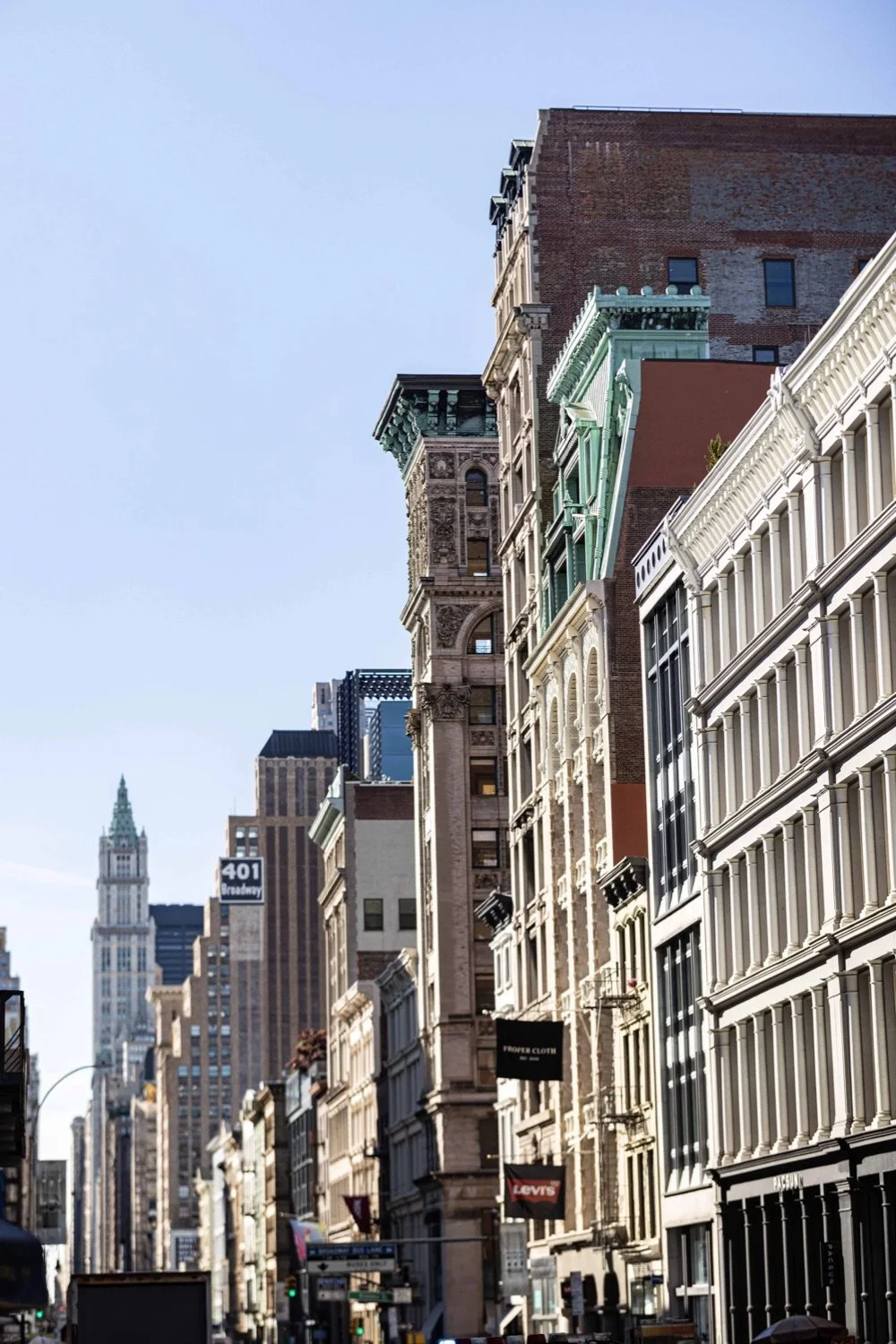 A city street view with tall historic and modern buildings under a clear blue sky. The buildings feature detailed facades, bay windows, and decorative elements, with some signs and street signals visible.