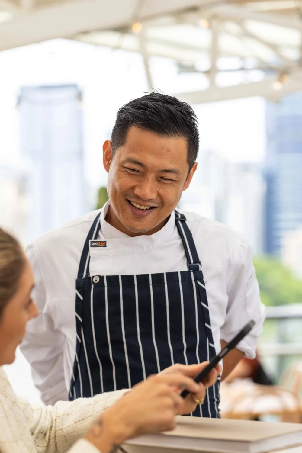 A smiling male chef wearing a striped apron and white chef's coat, talking to a woman who is holding a smartphone, in a bright restaurant or cafe setting.